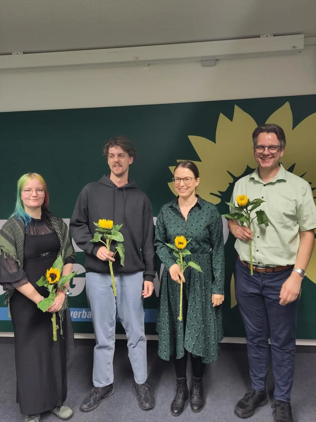 Die vier neu gewählten Vorstandsmitglieder stehen nebeneinander vor einer grünen Wand mit einer aufgemalten gelben Sonnenblume. Sie halten alle vier eine echte Sonnenblume in der Hand.