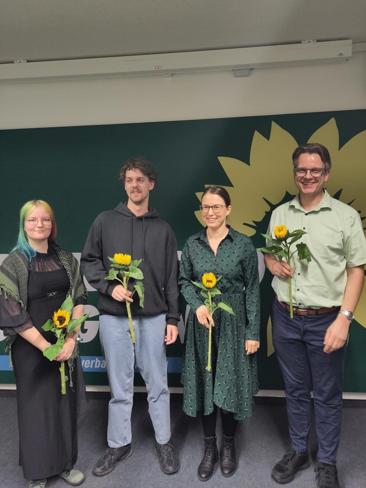 Die vier neu gewählten Vorstandsmitglieder stehen nebeneinander vor einer grünen Wand mit einer aufgemalten gelben Sonnenblume. Sie halten alle vier eine echte Sonnenblume in der Hand.